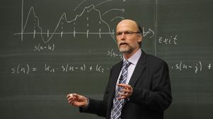 Dozent hält vor einer mit mathematischen Formeln beschriebenen Tafel einen Vortrag.