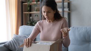 Unzufriedene Frau öffnet auf einem Sofa ein Paket und betrachtet den Inhalt.