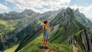 Wanderin steht auf einem Berggipfel und blickt über eine weite Gebirgslandschaft.