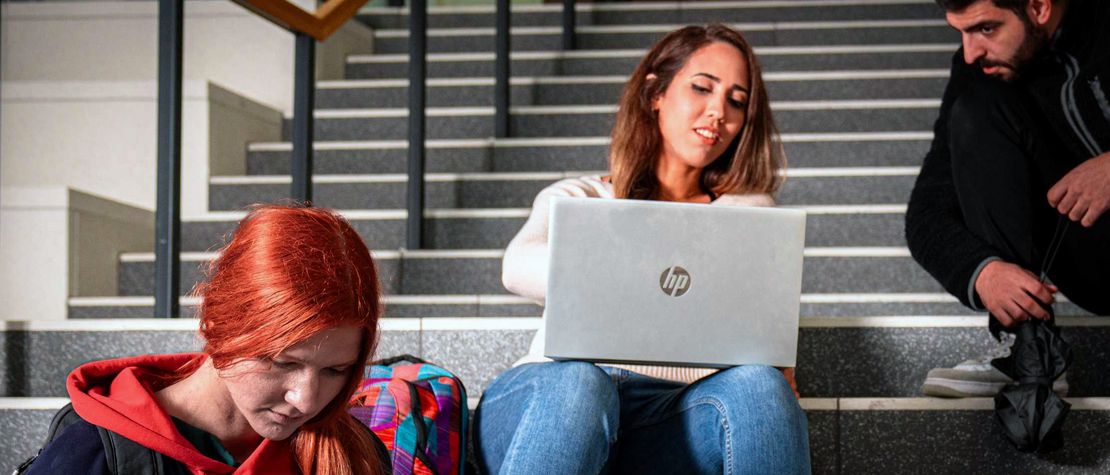 Zwei Studierende sitzen auf einer Treppe und arbeiten gemeinsam an einem Laptop.