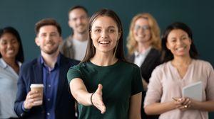 Mitarbeiterin reicht zur Begrüßung die Hand; hinter ihr steht ein diverses Team.