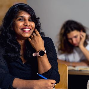 Lächelnde Studentin sitzt mit einem Stift in der Hand in einem Unterrichtsraum, im Hintergrund sind weitere Studierende zu sehen.