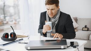 Erkrankter Mann sitzt mit Schal und einer Tasse vor einem Laptop.