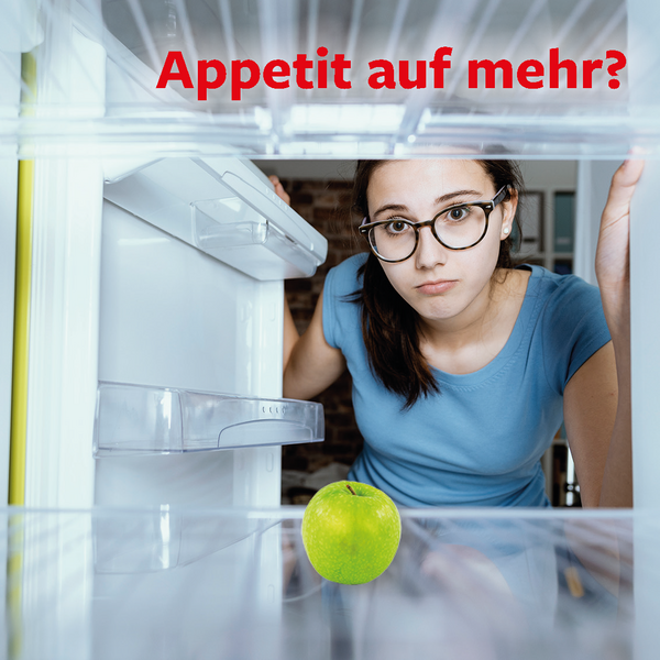 Eine junge Frau schaut hungrig in einen Kühlschrank, indem nur ein grüner Apfel liegt.