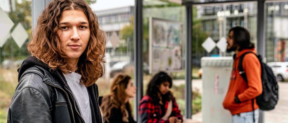 Junger Student mit langen Haaren steht mit weiteren Studierenden an einer Haltestelle.