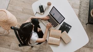 Studentin arbeitet an einem Laptop; neben ihr liegen mehrere aufgeschlagene Bücher. Ansicht von oben