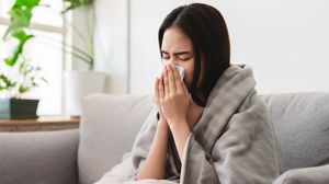 Erkältete Frau sitzt in eine Decke gehüllt auf einem Sofa und niest in ein Taschentuch.