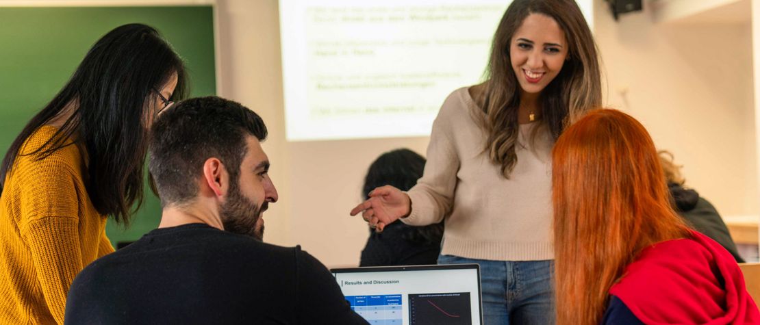 Studentin steht vor einer Lerngruppe und erklärt etwas; auf dem Tisch steht ein Laptop.