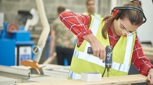 Handwerkerin mit Schutzbrille, Gehörschutz und Warnweste bohrt in eine Holzplatte.
