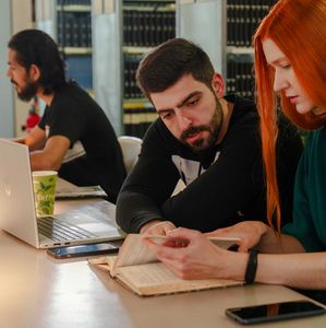 Zwei Studierende lesen und besprechen gemeinsam ein aufgeschlagenes Buch in einer Bibliothek.
