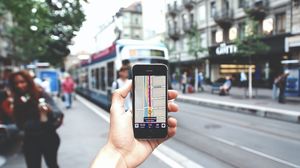 Hand hält ein Smartphone mit einer Navigationskarte vor einer Straßenbahnhaltestelle.